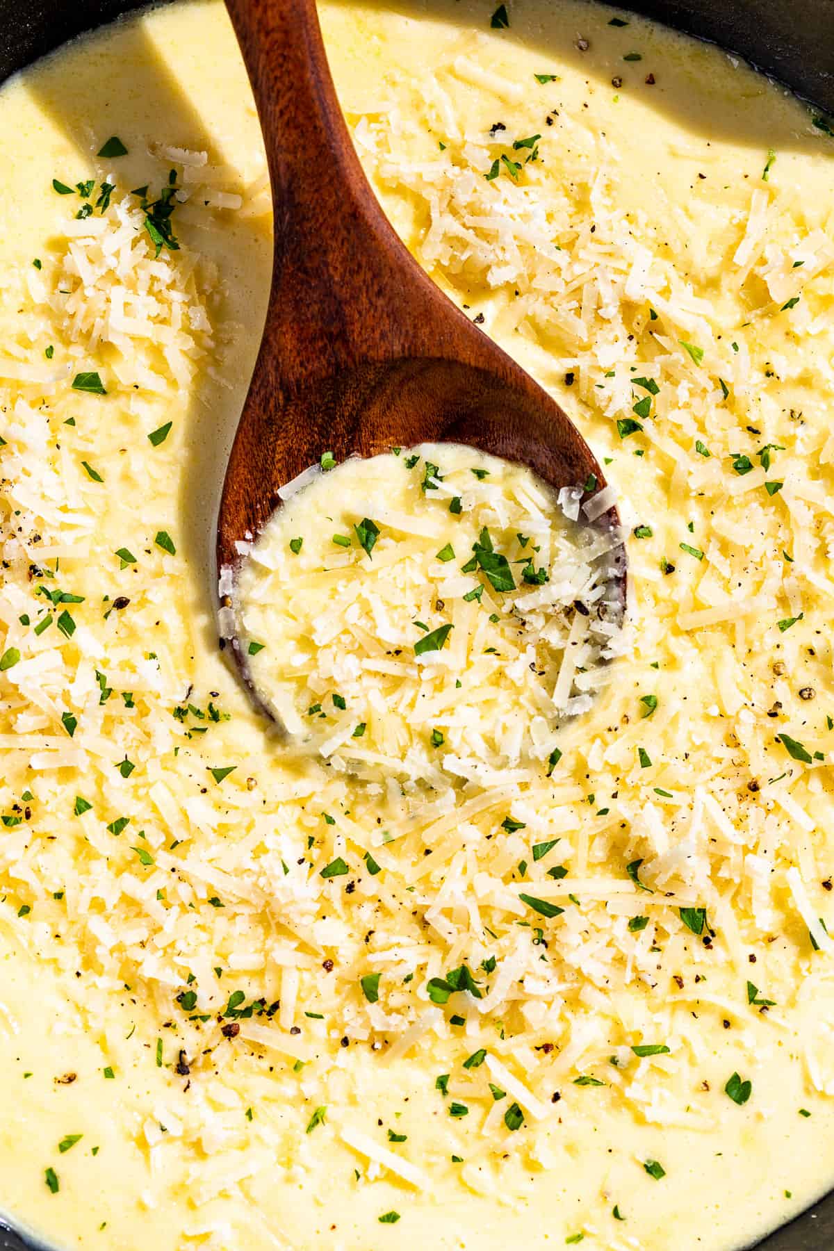 Close macro view of Garlic Parmesan Sauce topped with grated parmesan and sprinkled with chopped parsley with a wood spoon in it.