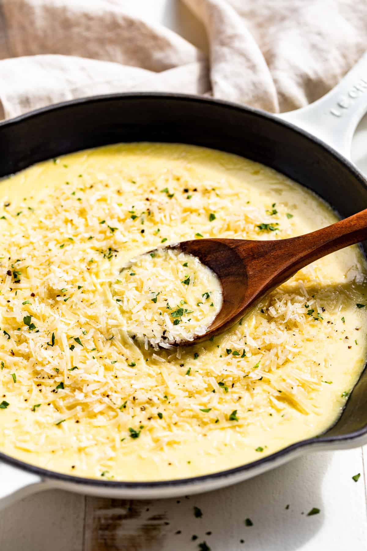 Side view of Garlic Parmesan Cream Sauce in a white enameled skillet topped with grated parmesan cheese, chopped parsley, and a wood spoon in the side of the dish.