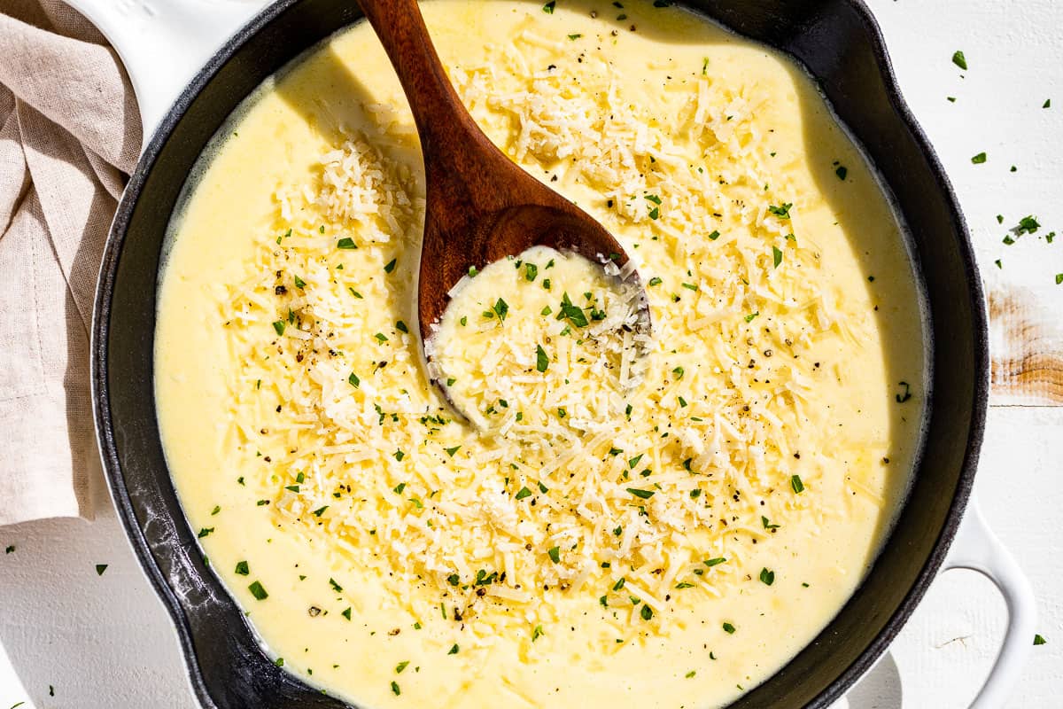 Finished Garlic Parmesan Sauce topped with grated parmesan and chopped parsley in a white enameled skillet with a wood spoon in it.