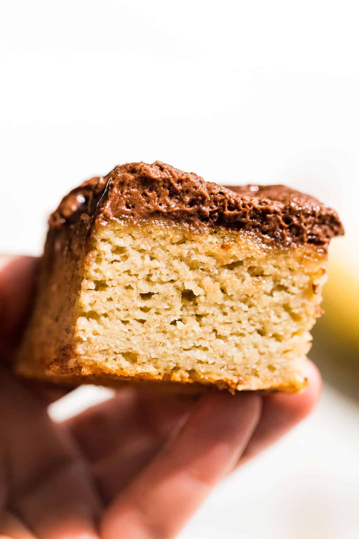 A hand holding a slice of gluten free banana cake topped with chocolate buttercream against a white background.