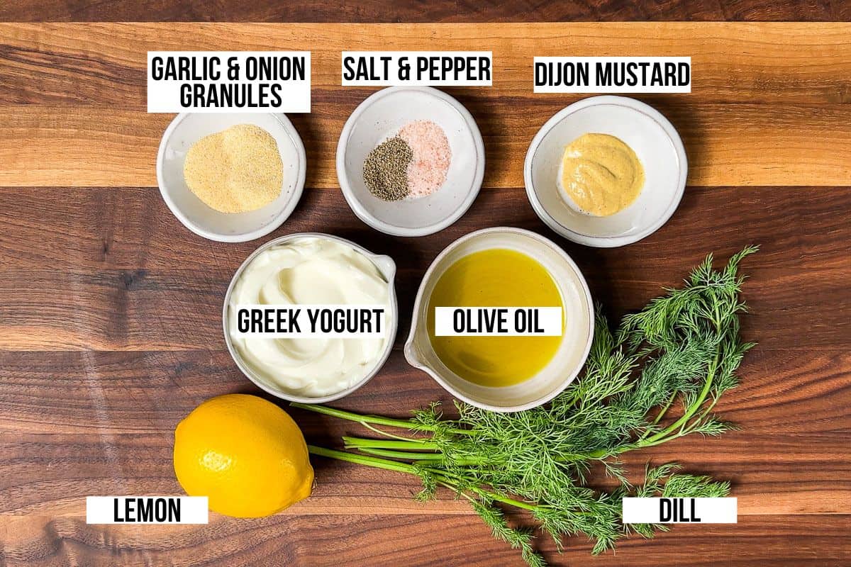 Greek yogurt, olive oil, Dijon mustard, dill, lemon, garlic and onion granules on a wood cutting board.