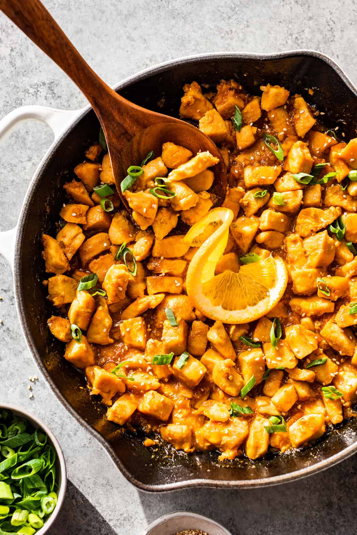 Straight down view of half a skillet of healthy orange chicken with a wood spoon scooping some out and sliced green onions on the side.