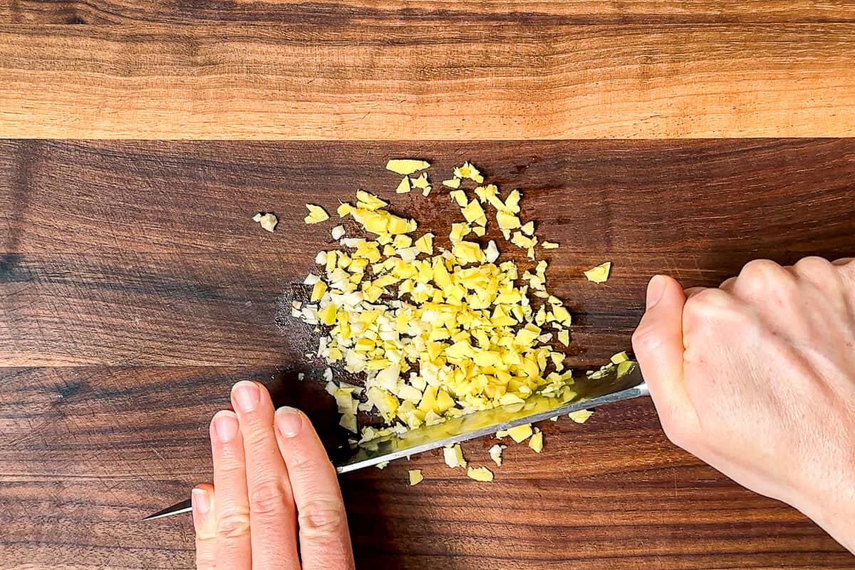 Mincing the garlic and ginger on a wood cutting board with a chef's knife.