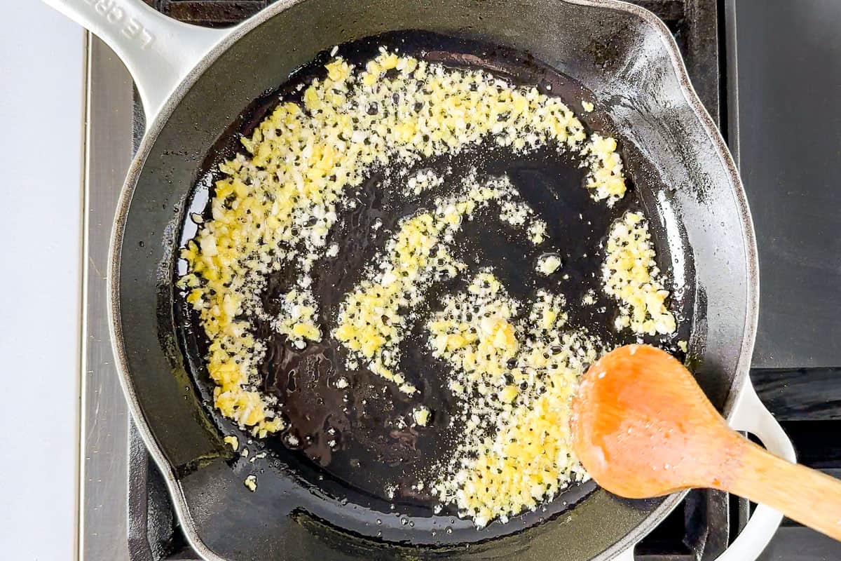 Sautéing the garlic and ginger in a white enameled cast iron skillet with a wooden spoon.