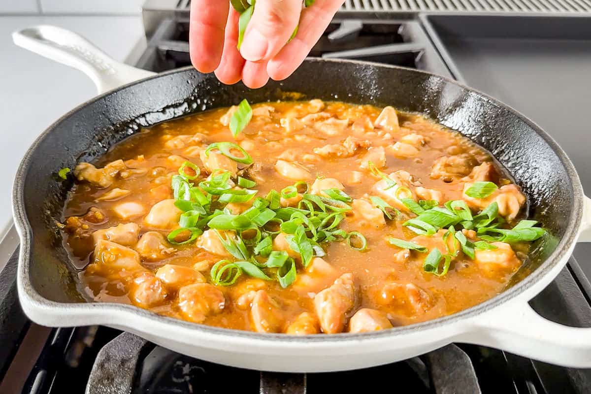 Sprinkling sliced green onions over the top of the finished healthy orange chicken in the skillet.