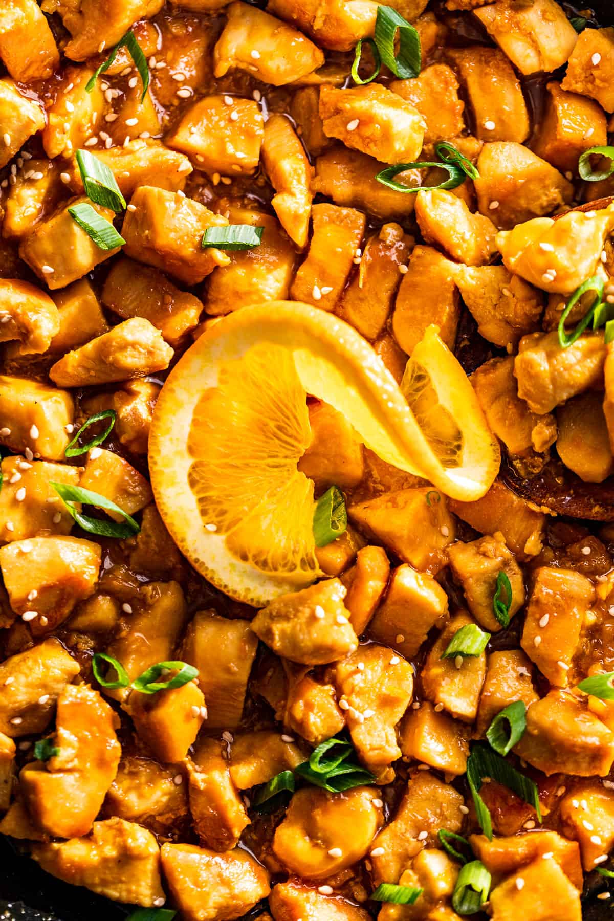 Close macro view of orange chicken topped with sliced green onions and a slice of an orange.