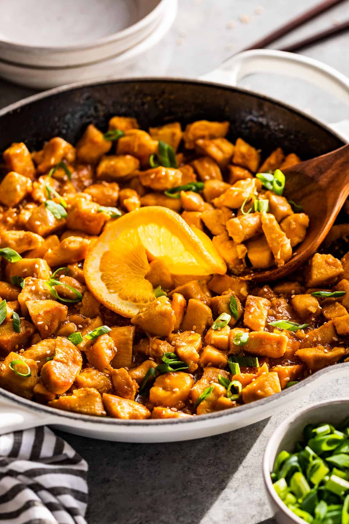 Healthy Orange Chicken in a white skillet with serving bowls, chop sticks, and sliced green onions!