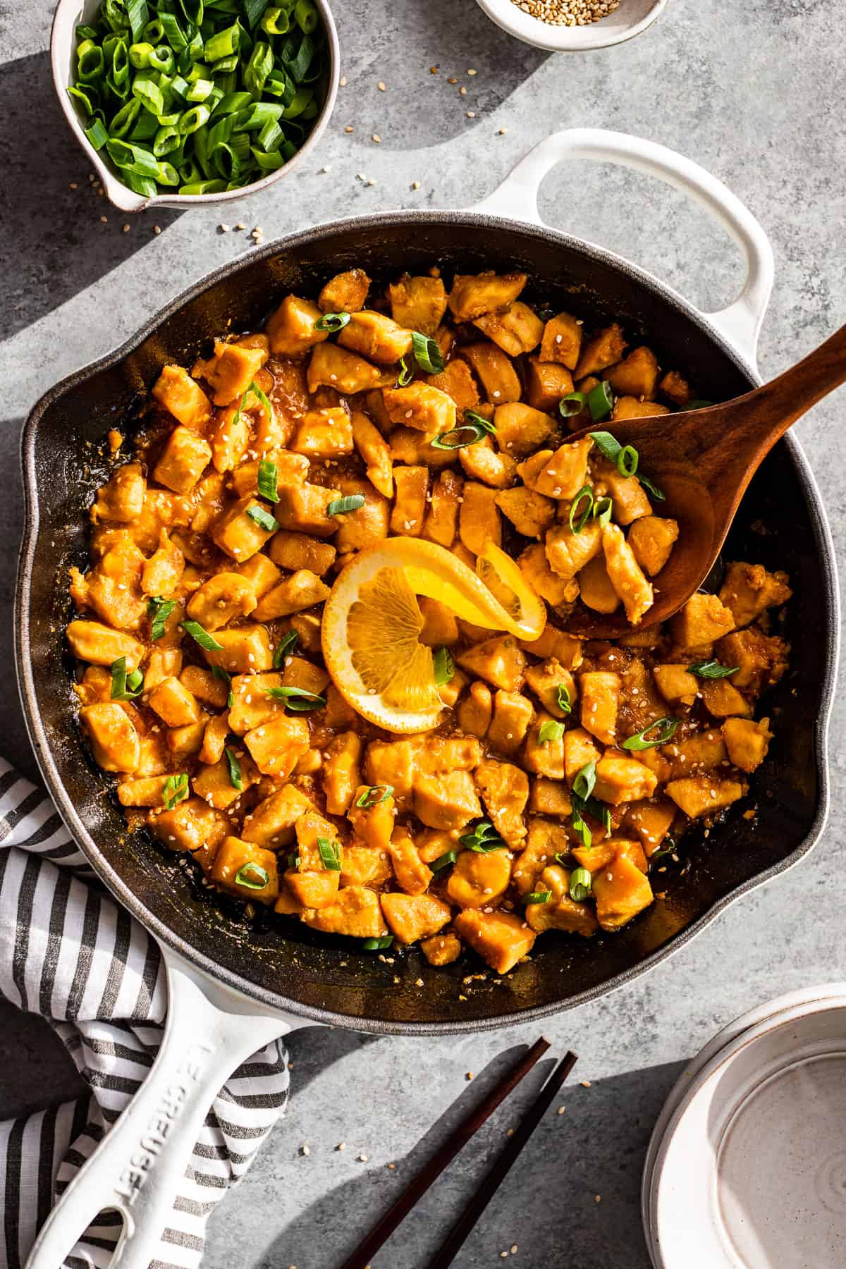 Straight down view of Healthy Orange Chicken in a white skillet with bowls of green onion and sesame seeds on the side.