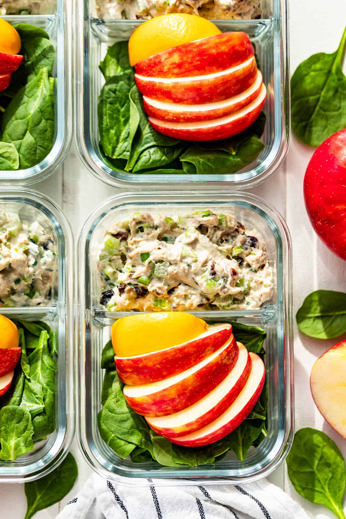 Downwards view of 4 meal prep containers filled with Healthy Tuna Salad, spinach, sliced apples, and a lemon wedge.
