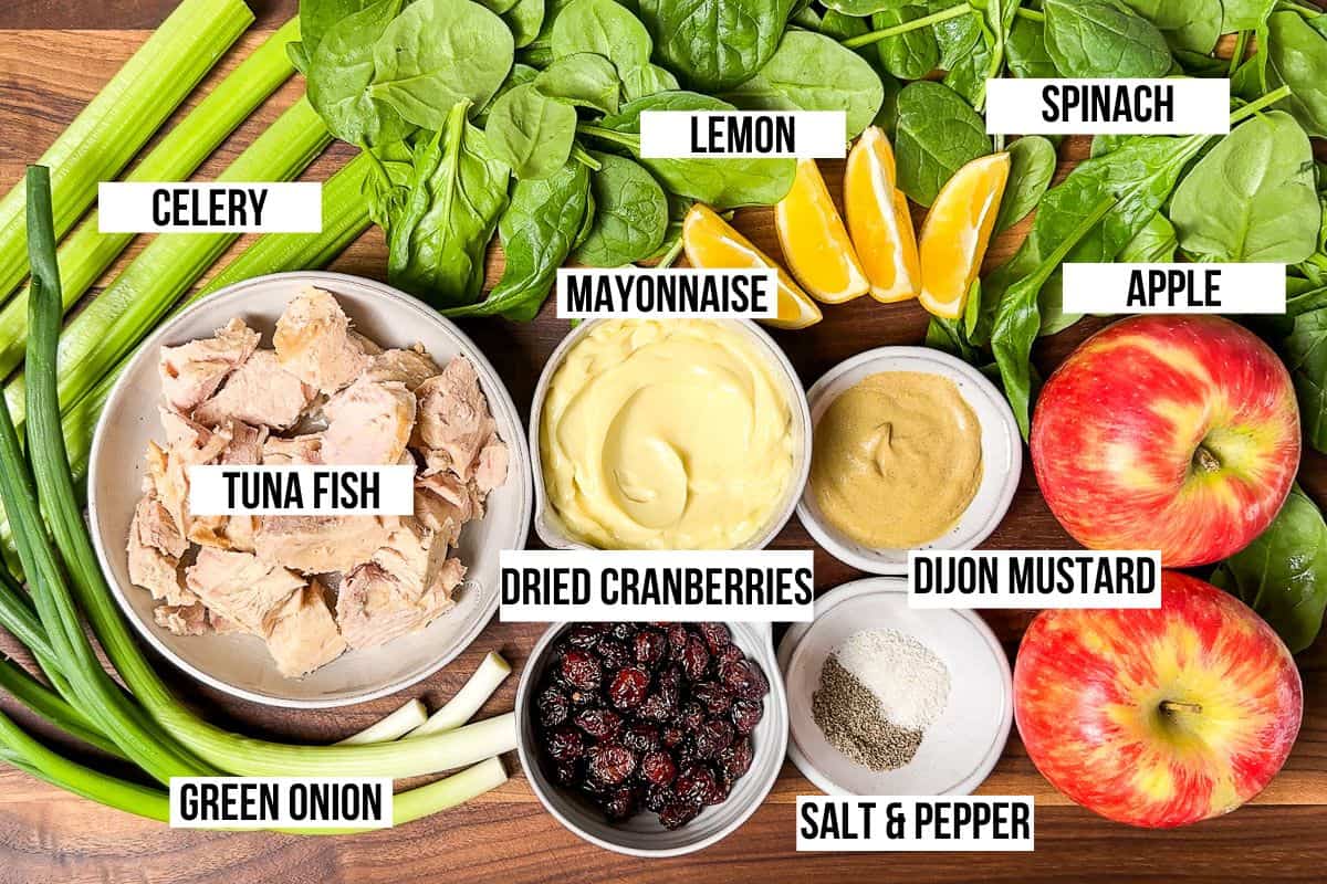 Canned tuna fish, green onion, celery, mayonnaise, apples, dried cranberries, lemon, spinach, and Dijon mustard in bowls on a wood cutting board.