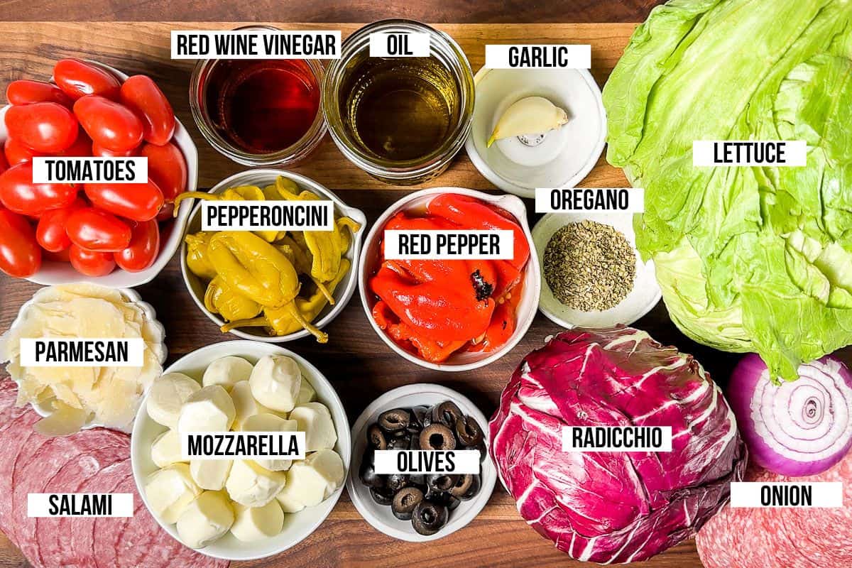 All the ingredients for Italian Chopped Salad on a wood cutting board: salami, mozzarella, provolone, olives, roasted red pepper, pepperoncini, radicchio, iceberg lettuce, cherry tomatoes, oil and vinegar.