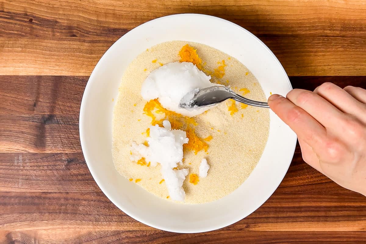 Adding the coconut oil to the lemon zest and sugar in a white bowl on a wood cutting board.