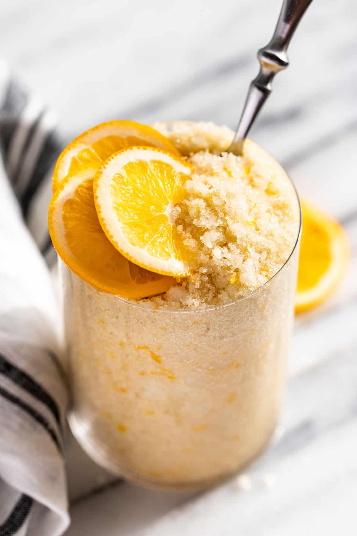 Side view of Lemon Sugar Scrub in a clear glass with lemon slices on top and a blue and white striped linen on the side.