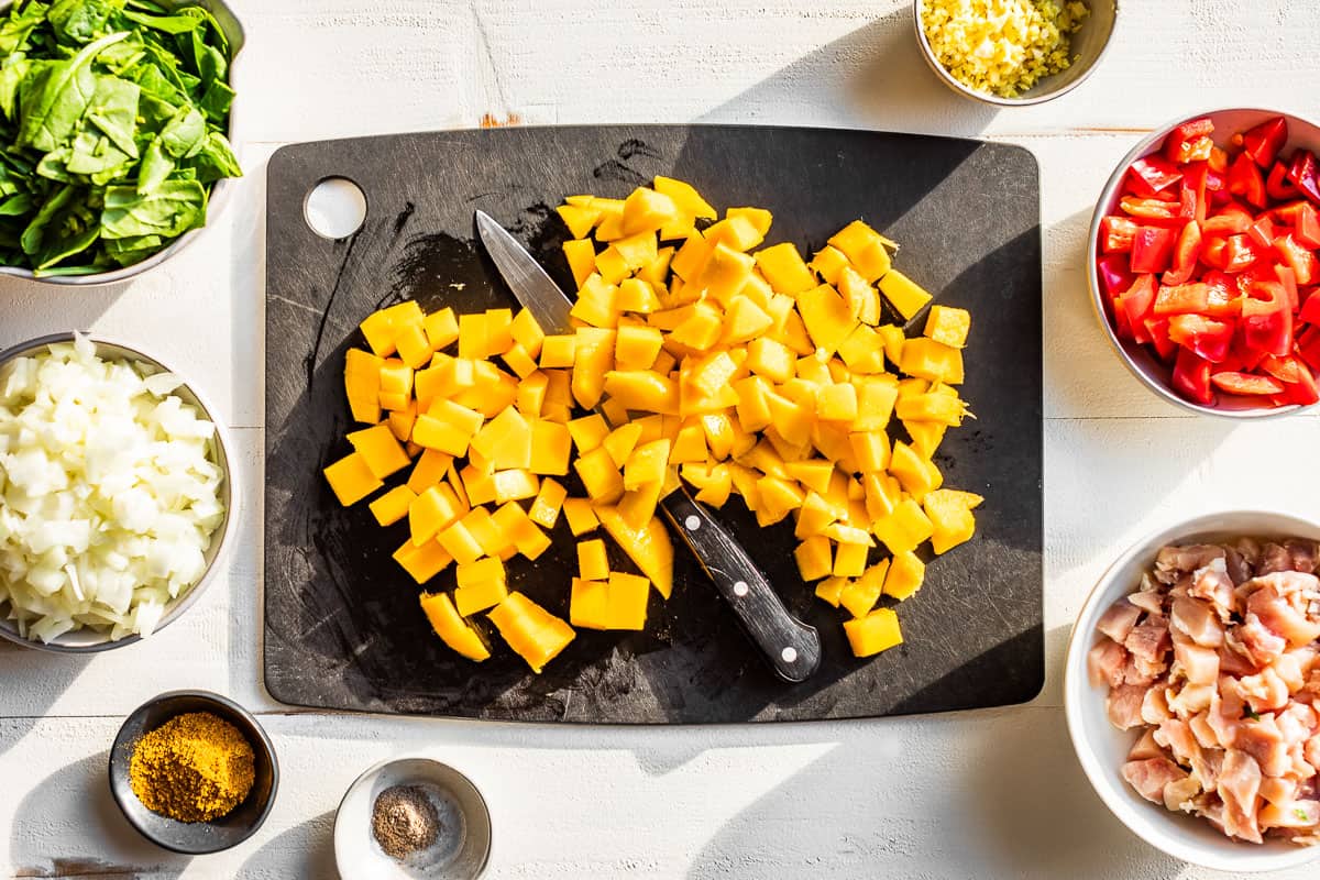 Chopped onion, minced ginger, diced chicken thighs, diced bell pepper and diced mango in bowls and a cutting board.