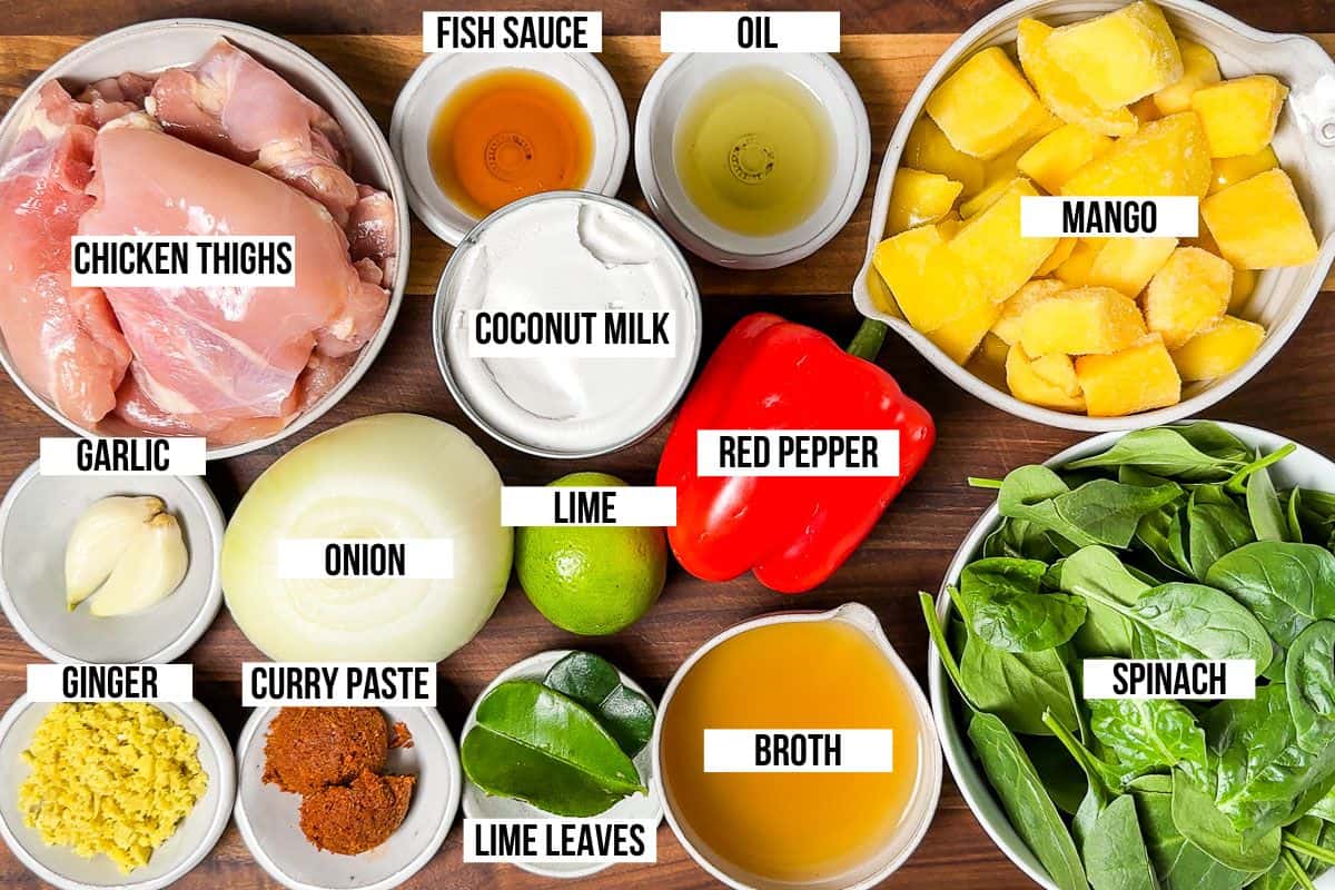 Chicken thighs, onion, garlic, ginger, curry paste, mango, spinach, bell pepper, coconut milk, lime, fish sauce, and broth on a wood cutting board.