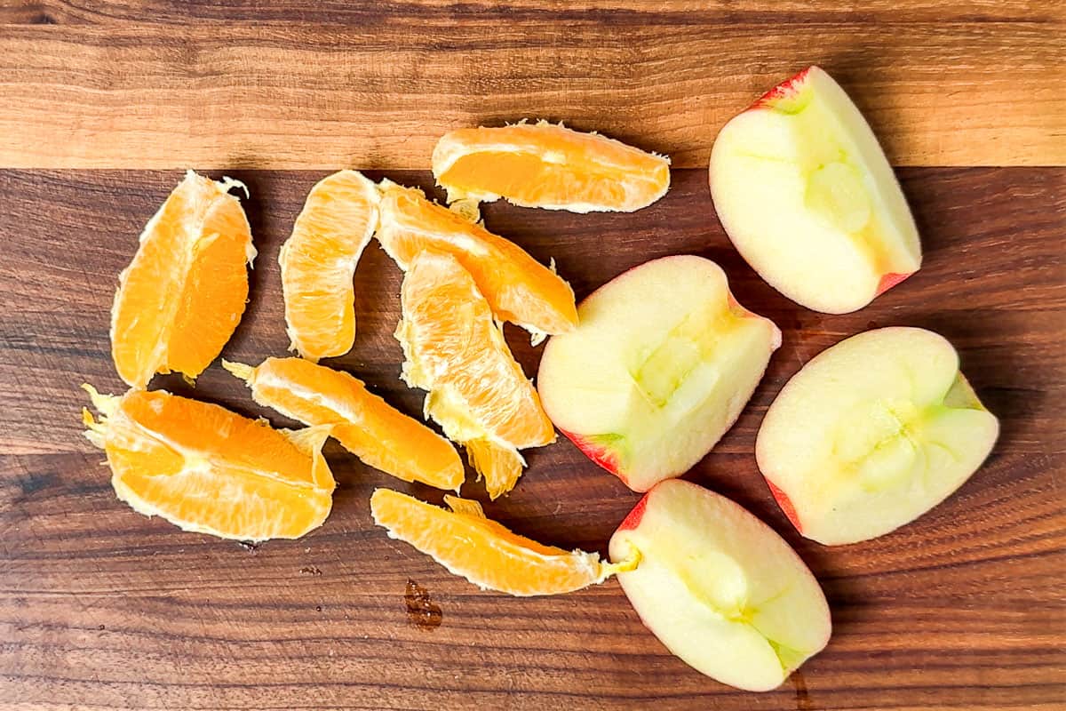 A quartered apple, and a peeled sectioned orange on a wood cutting board.