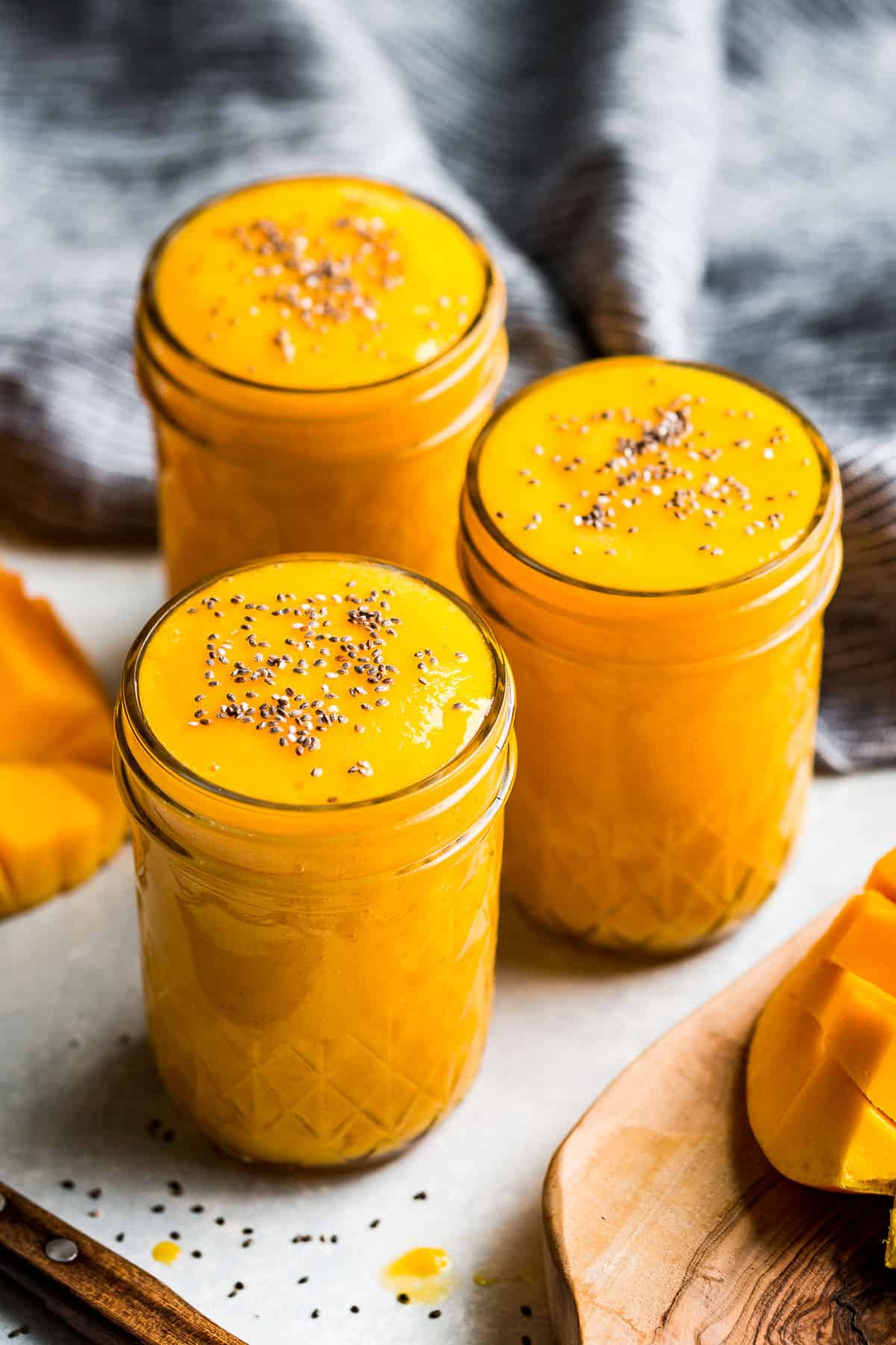 Side view of Mango Smoothie in three glasses sprinkled with chia seeds and a blue linen in the background.