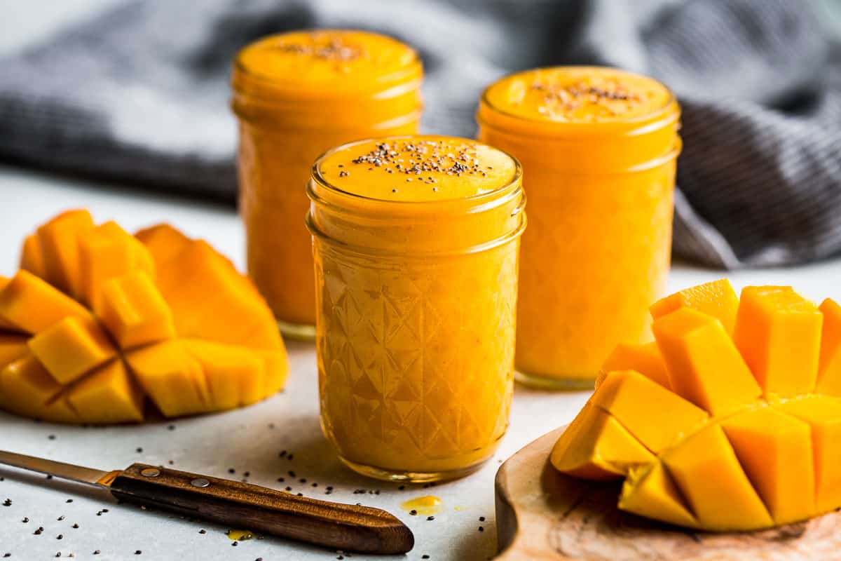 Three glasses of Mango Smoothie with two mango halves on the side and a blue linen in the background.