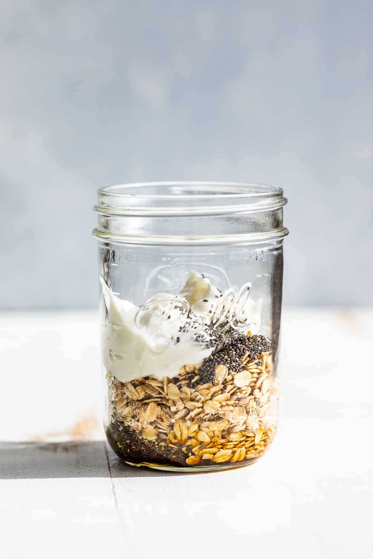 Oats, chia seeds, maple syrup, and yogurt added to a clear glass jar.