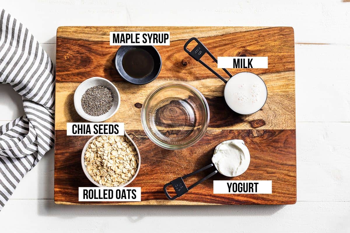 Oats, chia seeds, maple syrup, yogurt, milk and a clear glass jar on a wood cutting board.