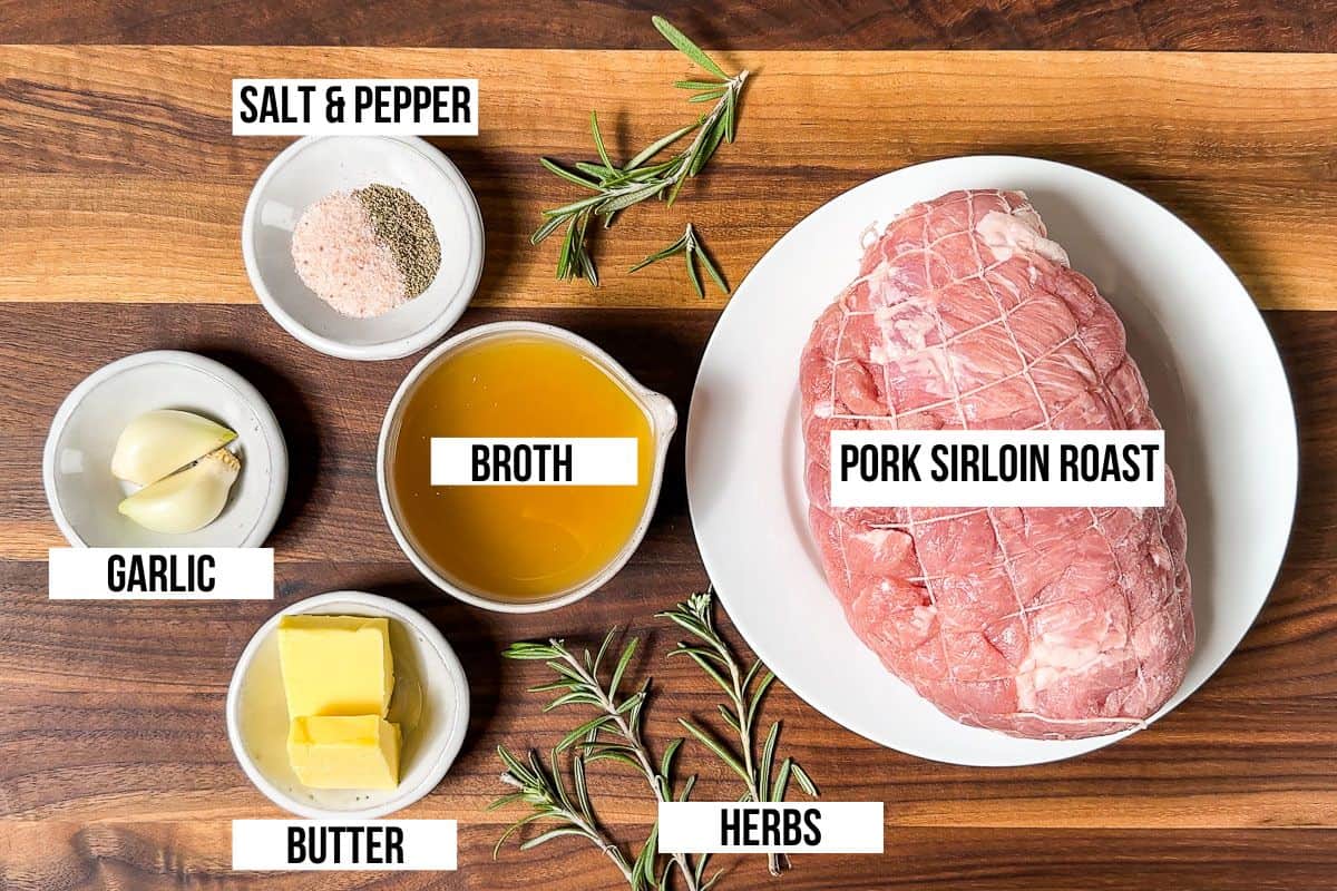 Pork sirloin roast, broth, butter, herbs, garlic, salt and pepper in dishes on a wood cutting board.