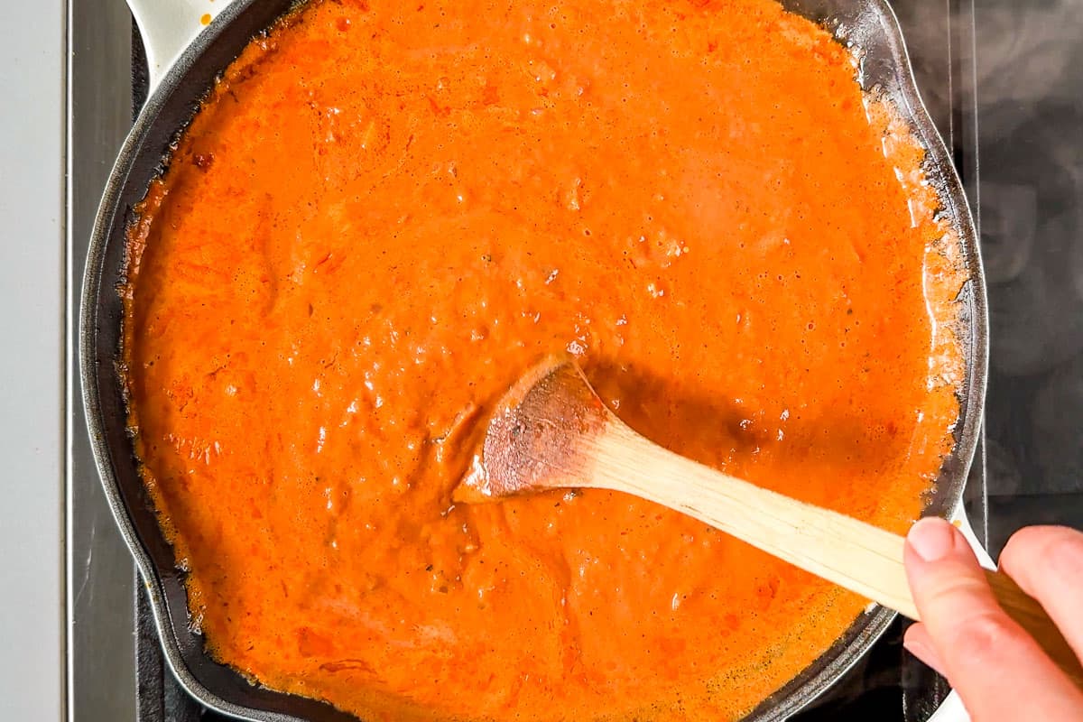 Finished Tomato Cream Sauce in a white cast iron skillet with a wood spoon stirring it.