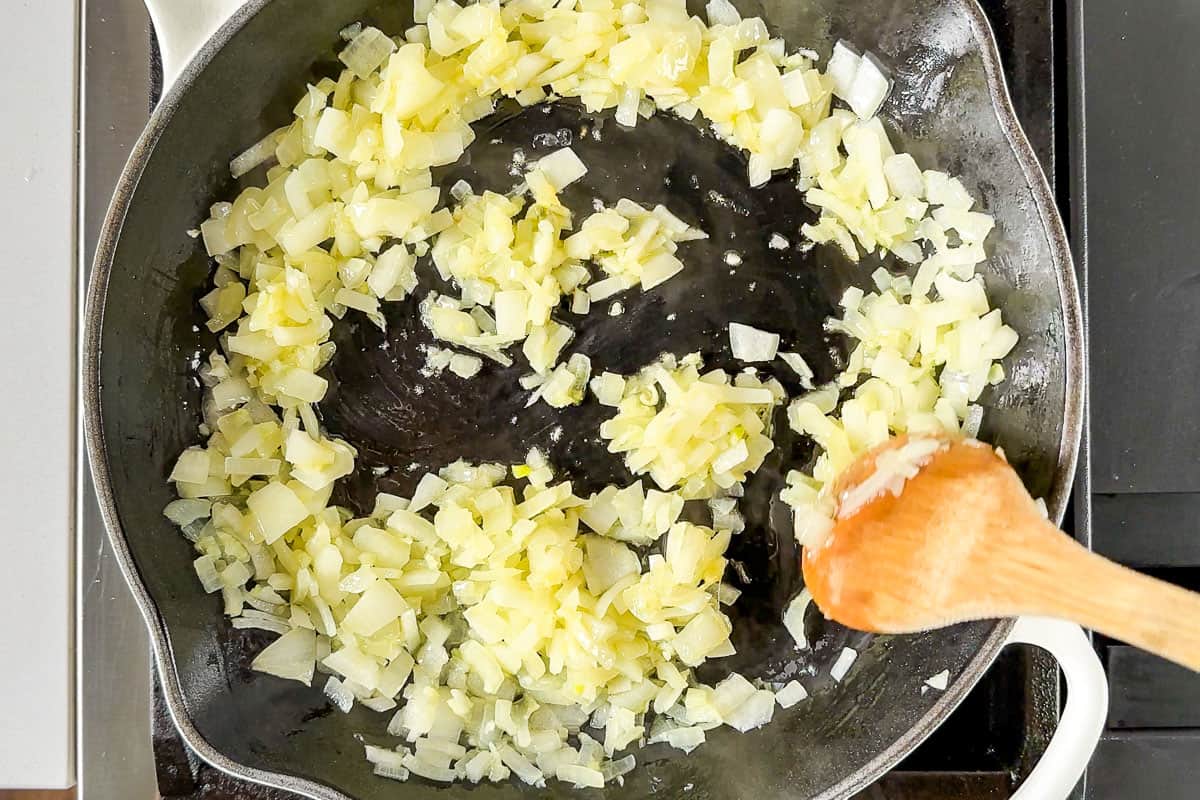 Sautéing onion and garlic in a white cast iron skillet on the stove top with a wood spoon.