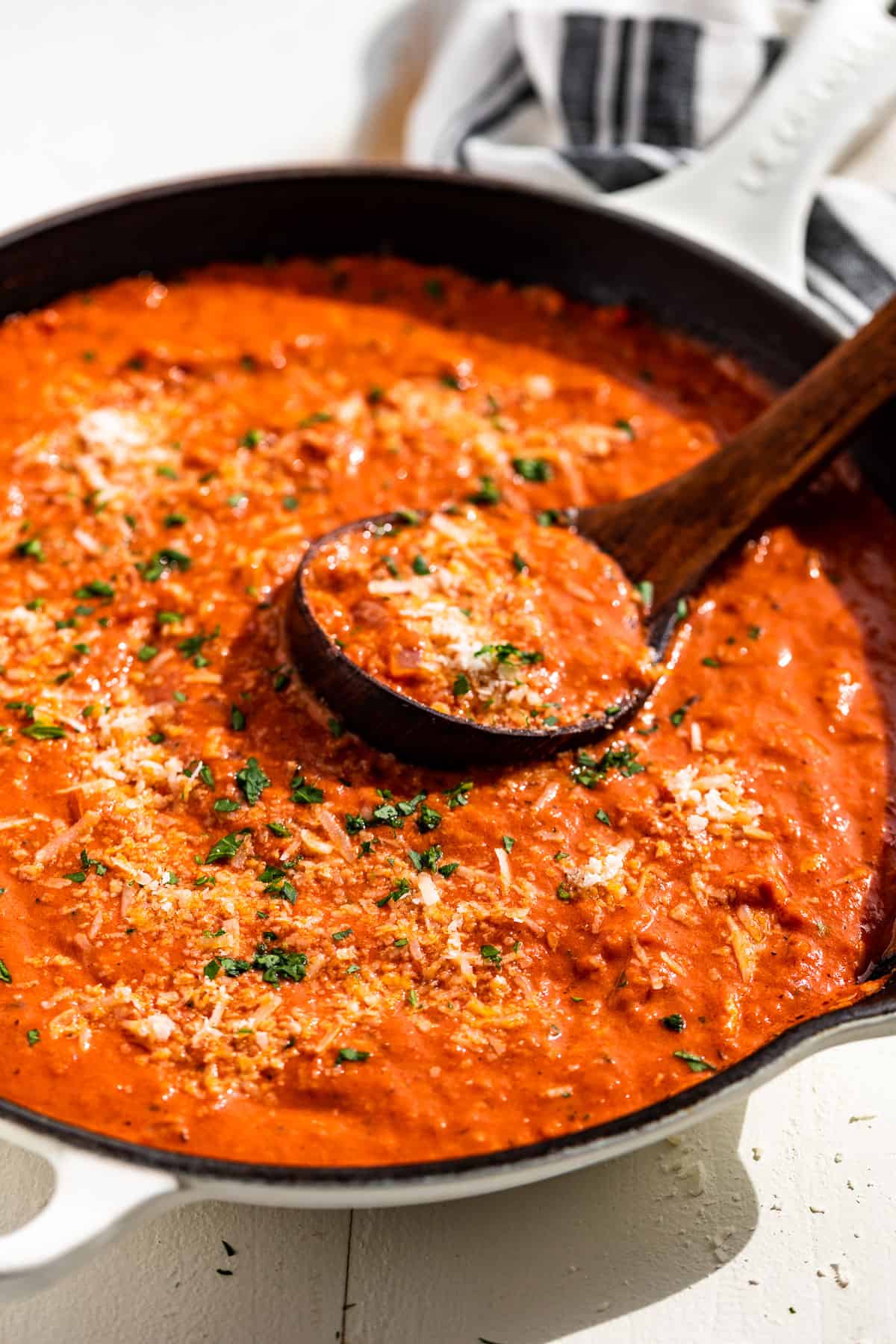 Side view of tomato cream sauce in a white cast iron skillet with a wood ladle in it and sprinkled with parmesan and parsley.