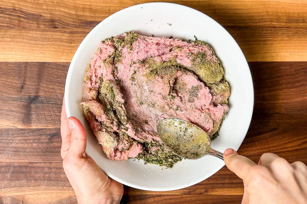 Mixing the herbs into the ground turkey with a metal spoon in a white bowl on a wood cutting board.