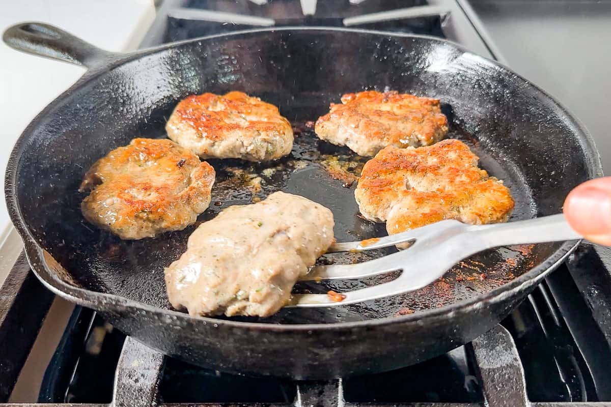 Pan frying the Turkey Breakfast Sausage patties in a cast iron skillet turning one over with a metal spatula.