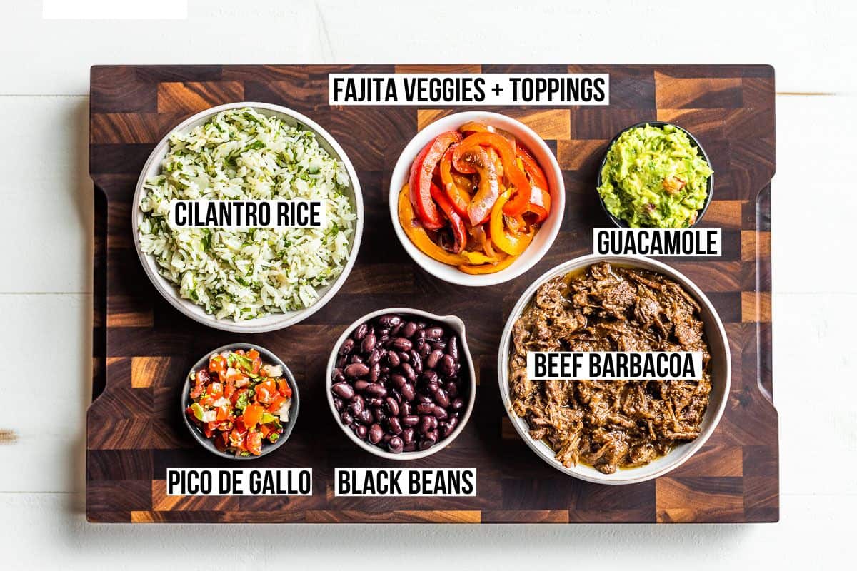 Bowls of cilantro lime rice, fajita veggies, shredded beef barbacoa, black beans, Pico de Gallo and guacamole on a wood cutting board.