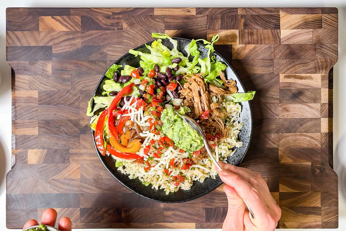 Topping the Carnitas Burrito Bowls with guacamole on a wood cutting board.