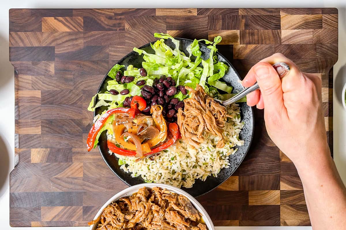 Adding carnitas, Fajita veggies, and black bean to the burrito bowl on a wood cutting board.