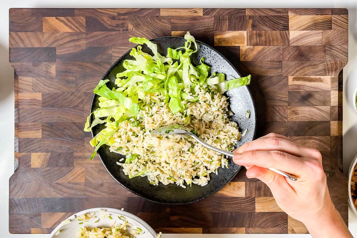 Adding cut up romaine and cilantro lime rice to a bowl on a wood cutting board.