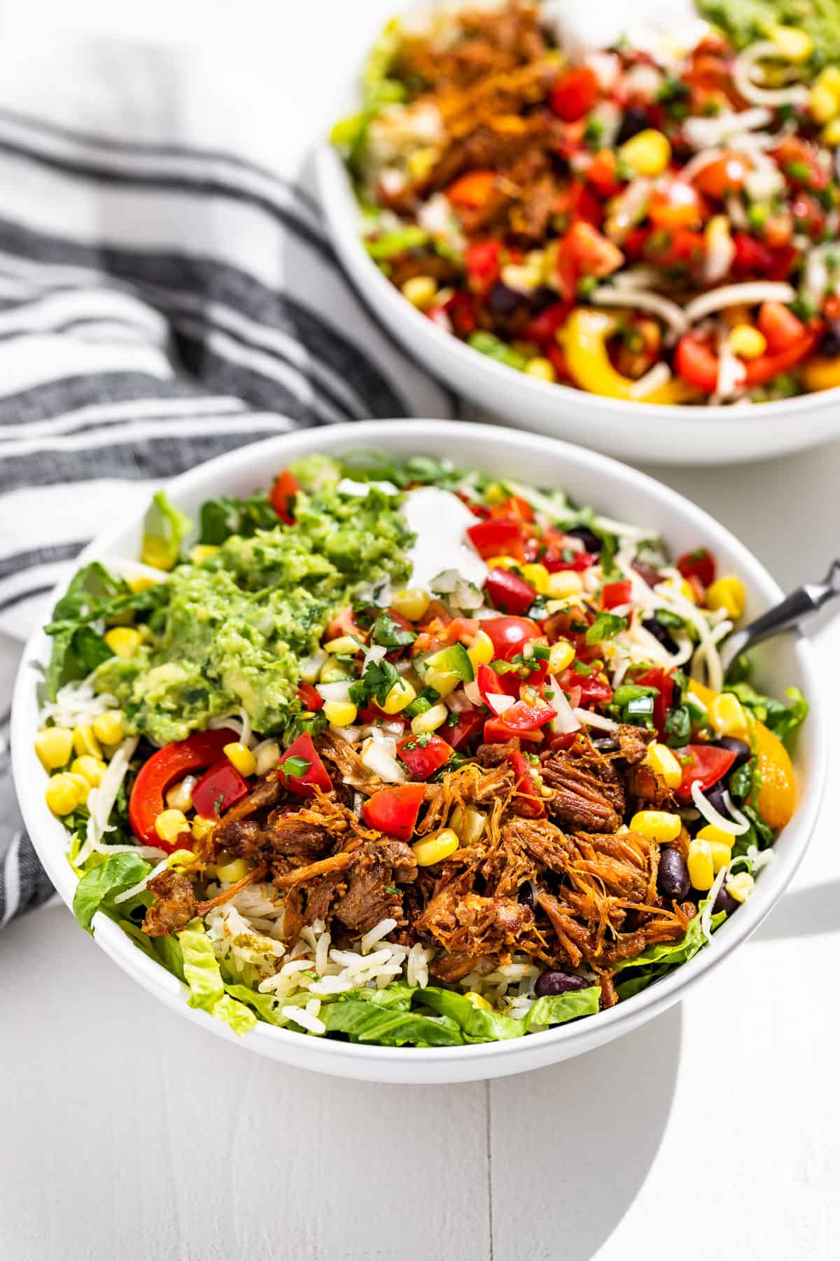 Side view of 2 Carnitas Burrito Bowls with forks on the side and blue and white striped linen on the side.