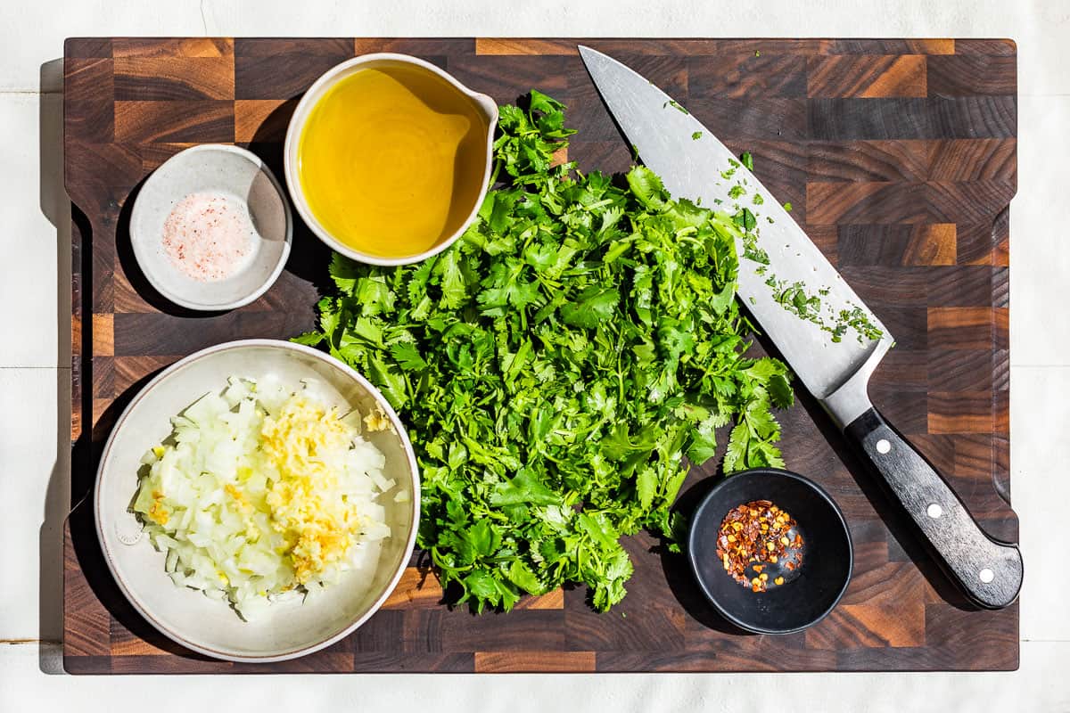 Diced onion, and chopped cilantro on a wood cutting board with small bowls of olive oil, salt, and red pepper flakes.