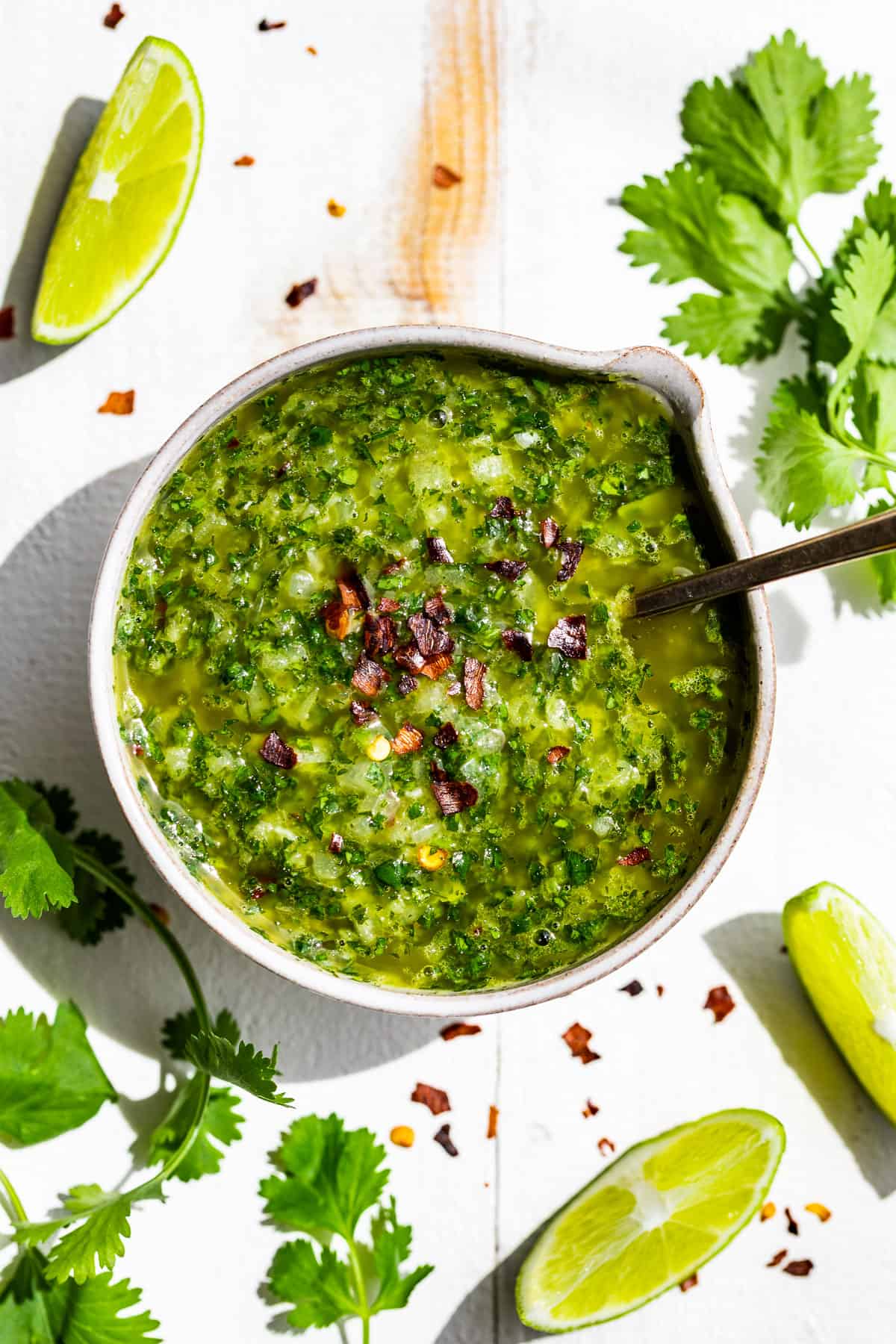 Cilantro Chimichurri in a pottery bowl on a white background surrounded by cilantro sprigs and lime wedges.