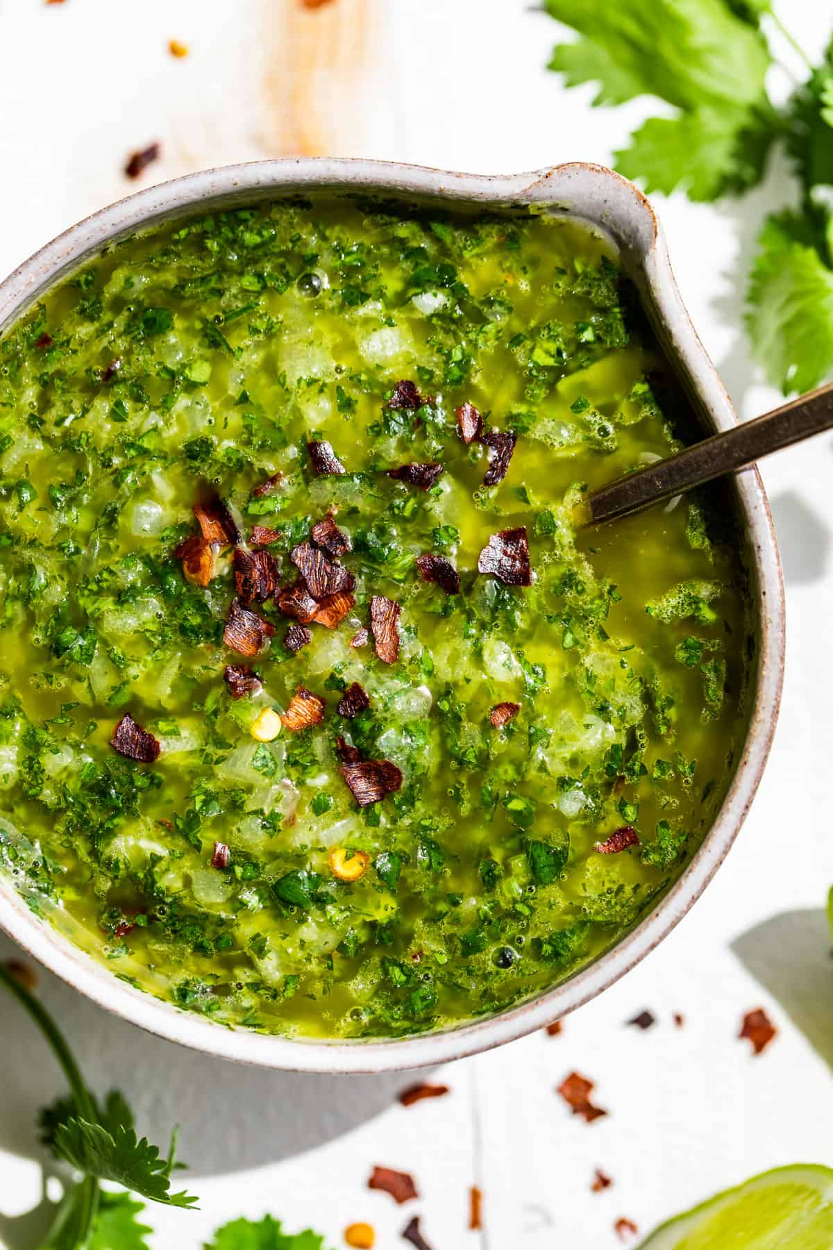 Straight down view of Cilantro Chimichurri in a pottery bowl with a silver spoon with cilantro sprigs and lime wedges around it.