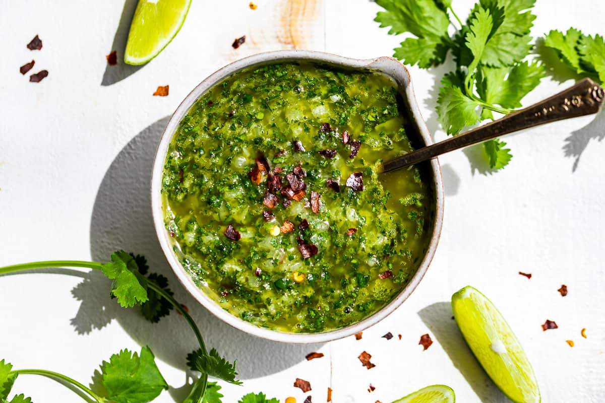 Straight down view of Cilantro Chimichurri in a small pottery bowl with a silver spoon in it and cilantro sprigs on the side.