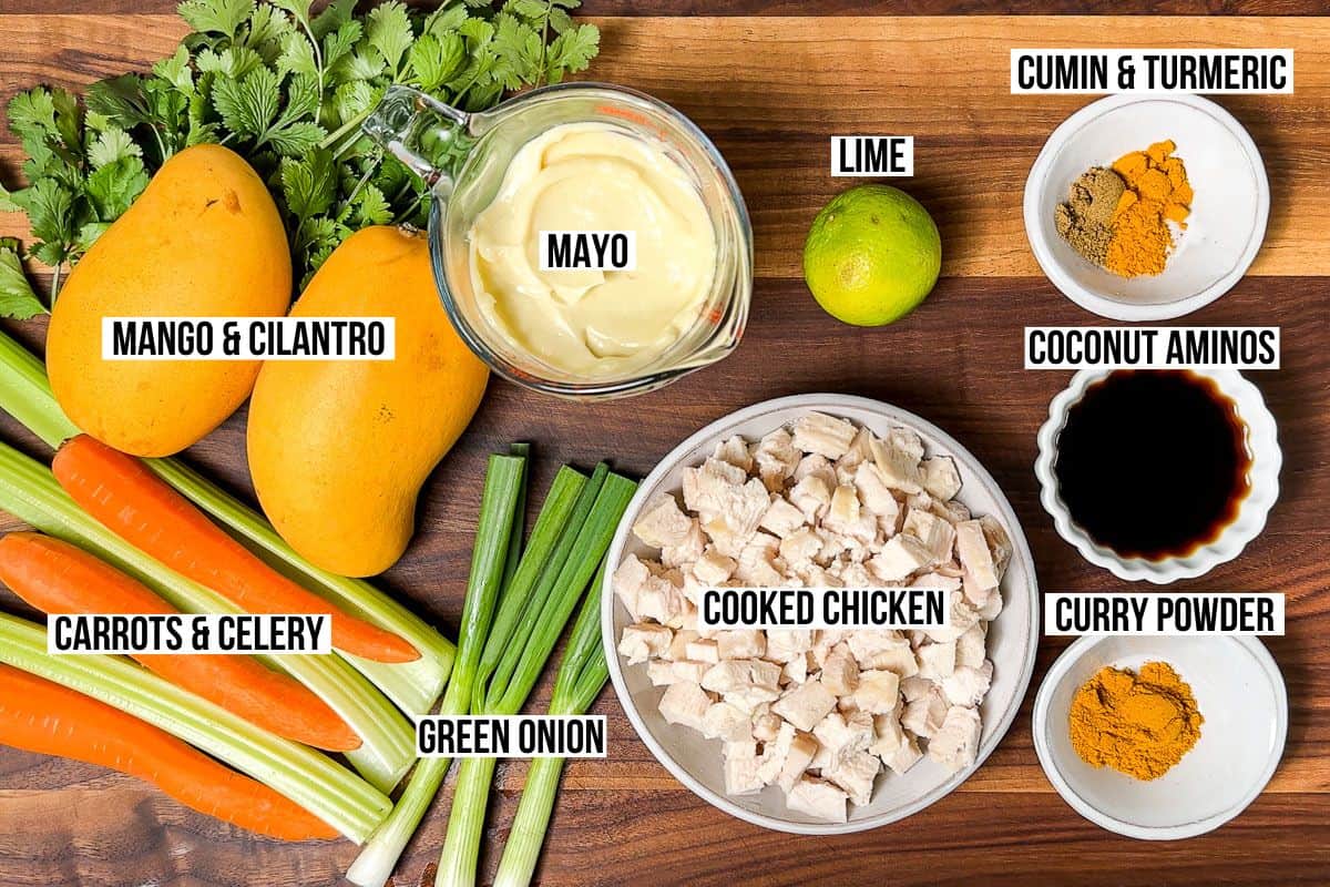 Diced cooked chicken, curry powder, green onion, mango, cilantro, lime, mayo, carrots, celery, and coconut aminos in small bowls on a wood cutting board.