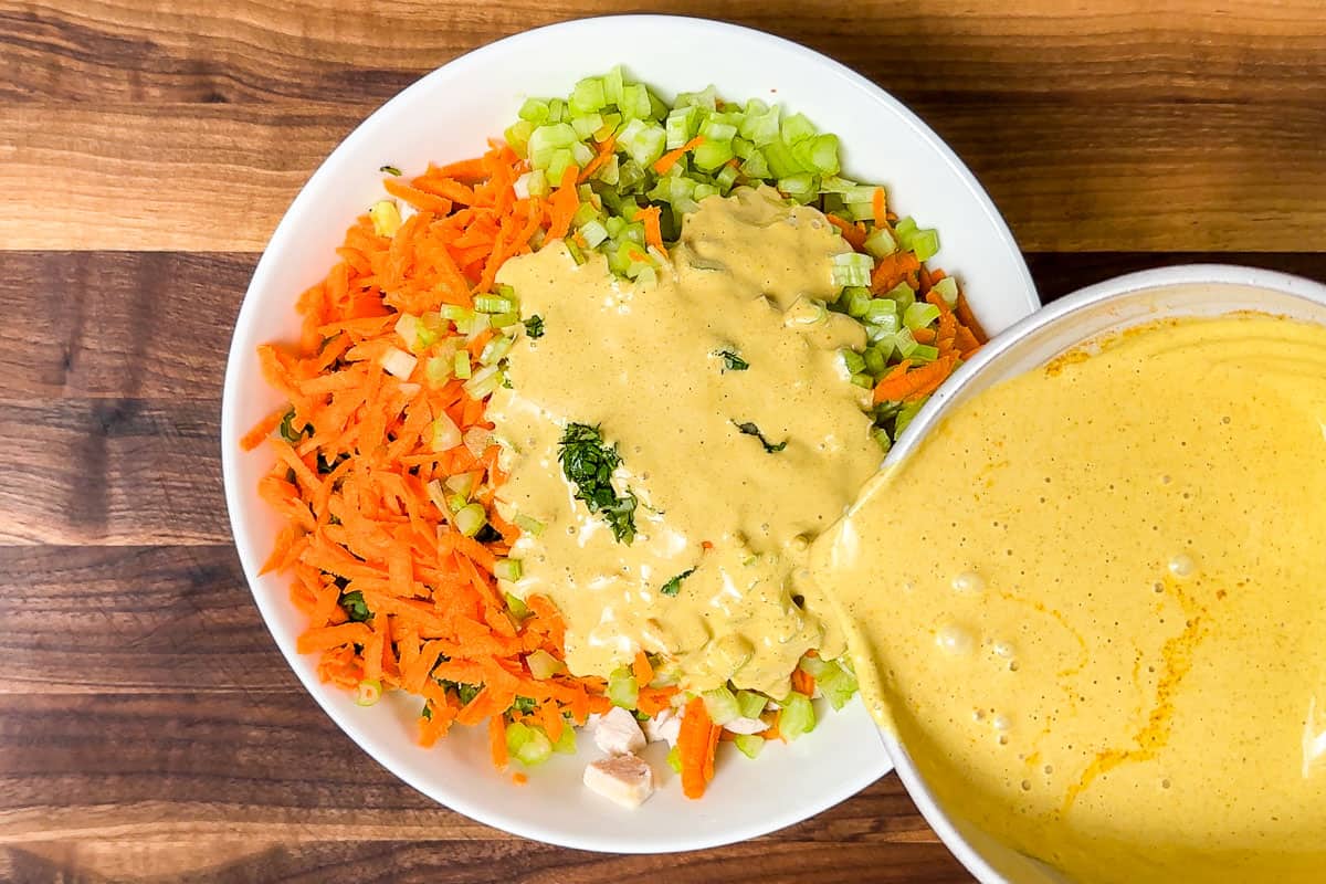 Pouring the curried mayo dressing over the carrots, celery, green onions, and chicken in a large white bowl.