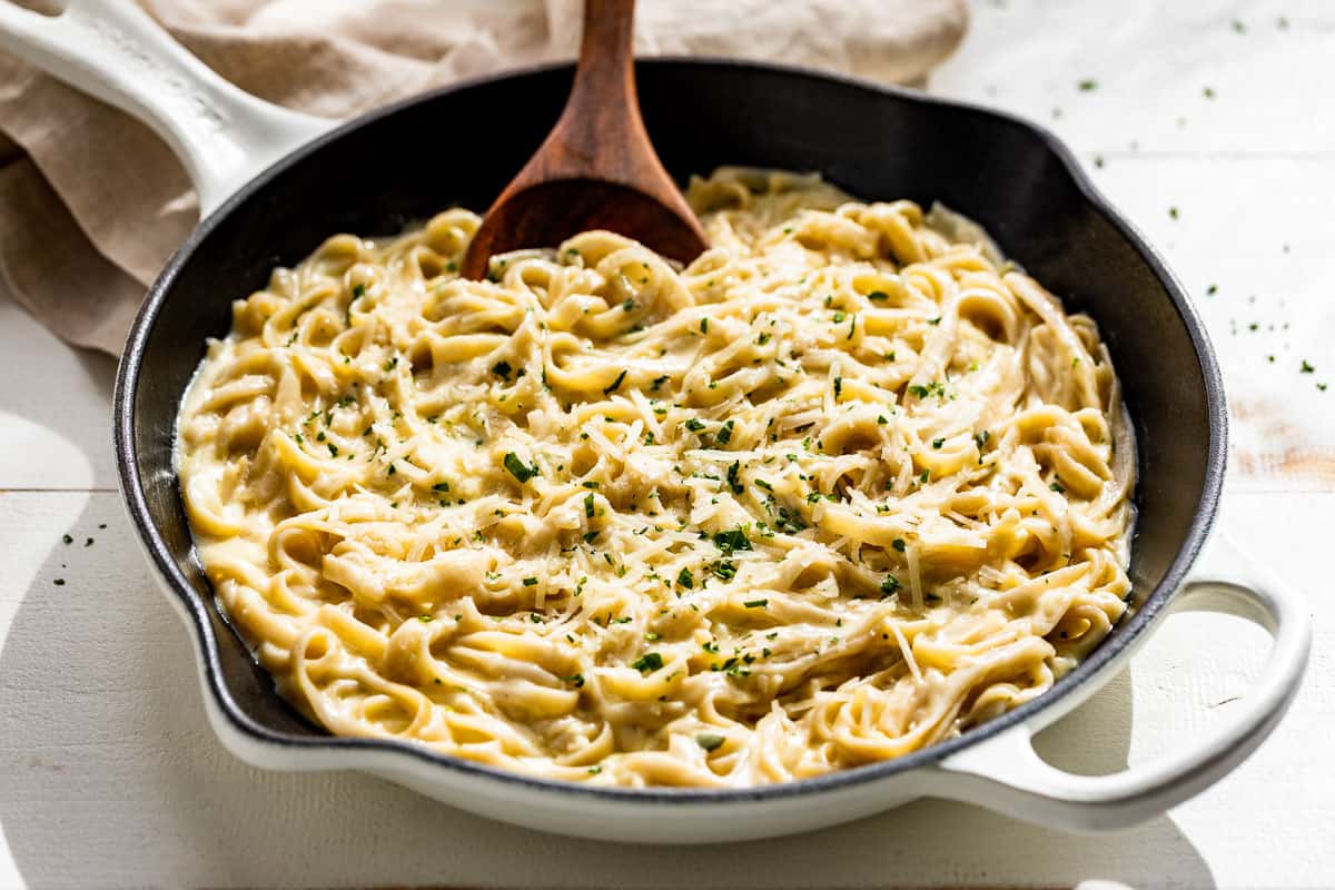 Side view of Garlic Parmesan Pasta sprinkled with chopped fresh parsley in a large white skillet with a wood spoon in it.