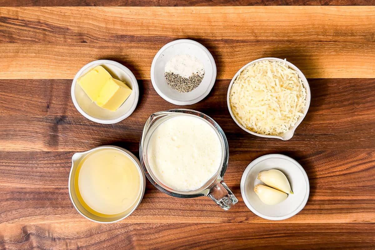 Heavy whipping cream, grated parmesan cheese, butter, garlic, white wine, sea salt and pepper in bowls on a wood cutting board.