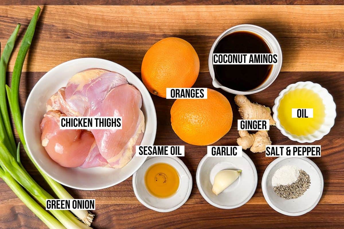 Chicken thighs, oranges, coconut aminos, ginger, garlic, sesame oil, oil, and green onions on a wood cutting board.