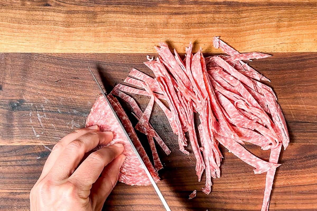 Thinly slicing the salami on a wood cutting board with a chef's knife.