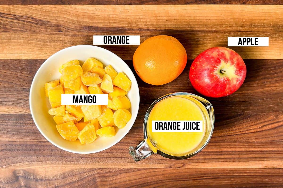 Frozen mango, an orange and apple, and orange juice in a glass on a wood cutting board.