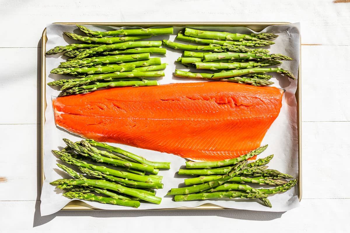Placing the salmon fillet on a parchment lined baking sheet with asparagus on the side.