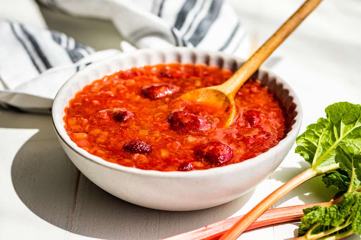 Strawberry Rhubarb Sauce in a grey pottery bowl with a wooden spoon with rhubarb stalks on the side with a blue and white linen on the other side.