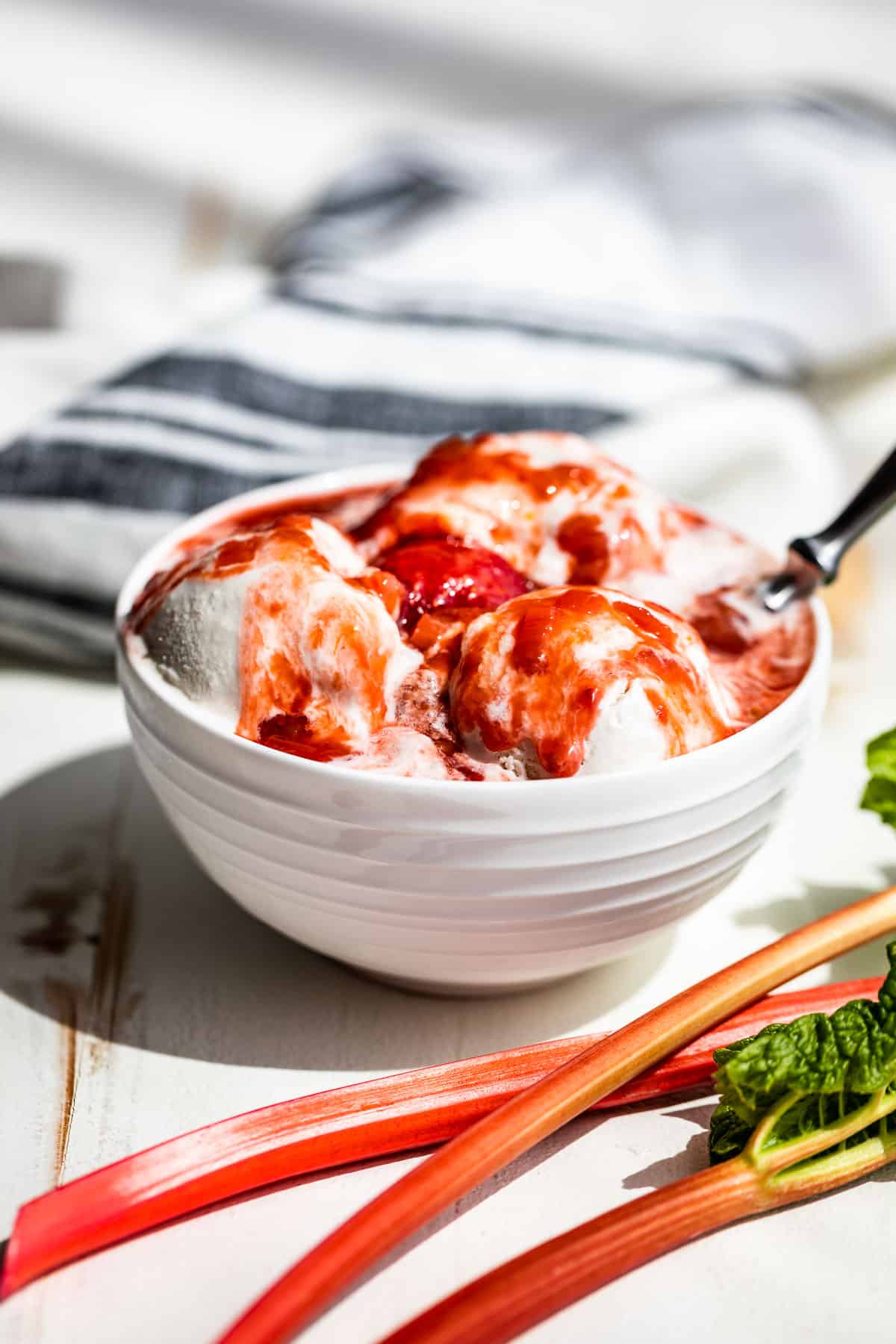 A white bowl with coconut ice cream topped with Strawberry Rhubarb Sauce with 3 stalks of rhubarb in the front and a blue and white linen in the background.
