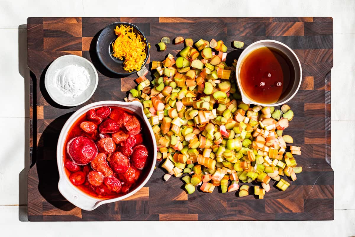 Sliced rhubarb, thawed frozen strawberries in a bowl, maple syrup, and orange zest in containers on a wood cutting board.